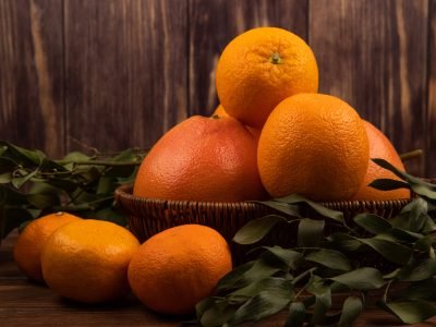 side view of fresh ripe oranges in a wicker basket and green leaves on dark wooden background